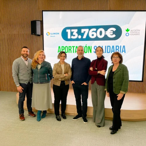 Six people pose in front of a screen displaying a €13,760 charitable donation from ILUNION Hotels to the organization 'Action Against Hunger'. The setting is modern, featuring a wooden backdrop and a circular platform.