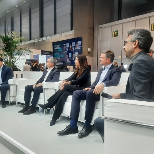 A group of people seated on white chairs. A large screen in the background displays their names along with logos including Barceló, Meliá, NH Hotel Group, and Palladium Hotel Group. The individuals are dressed in business attire.
