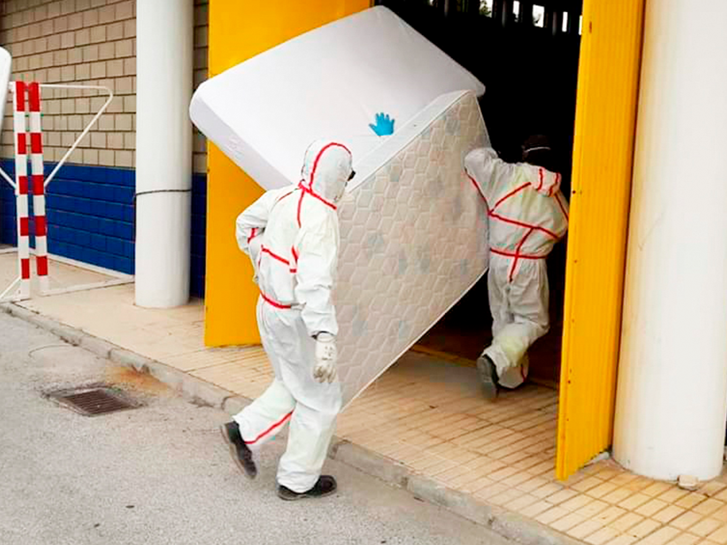 Dos personas con trajes de protección blanca con cintas rojas, además de guantes y botas negras. Están trasladando un colchón grande a través de una puerta amarilla.