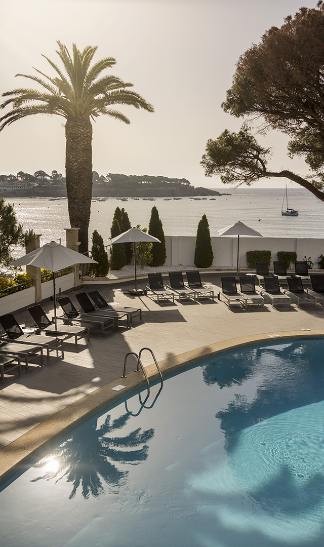 Piscina de un hotel al aire libre con tumbonas y sombrillas, rodeada de árboles, frente al mar.