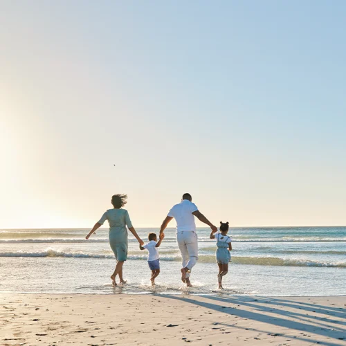Una familia de cuatro —dos adultos y dos niños— camina de la mano por la playa hacia el mar, iluminados por la luz suave del atardecer.
