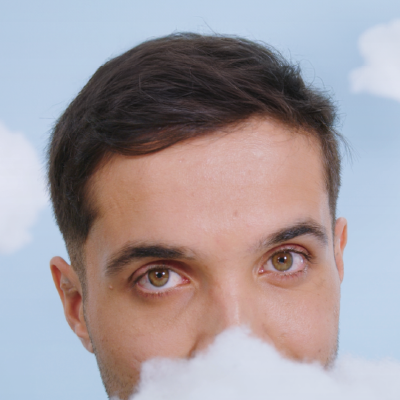 This image features a face partially covered by clouds, focusing on the eyes of a man. He has short, dark hair, set against a light blue background adorned with fluffy clouds, evoking a sense of lightness or dreaminess. His expressive eyes stand out due to their striking color, possibly symbolizing introspection or a connection to dreamlike and reflective themes. The framing suggests an artistic or conceptual visual message, perhaps related to dreams, imagination, or freedom.