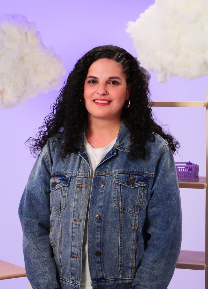 Woman with long, curly hair, wearing a blue denim jacket and a white T-shirt. The background is a soft shade of purple with a few scattered white clouds, evoking a dreamlike or introspective atmosphere.