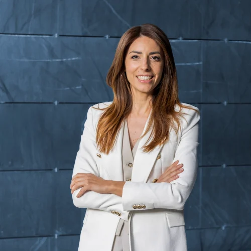 Beatriz Miguel, Business Director at ILUNION Hotels. A smiling woman with long brown hair, wearing a white blazer and standing with her arms crossed. The background features dark textured panels, conveying professionalism and confidence.