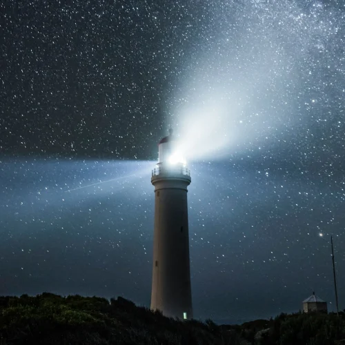 A starry night landscape illuminated by the light of a lighthouse on a hill.