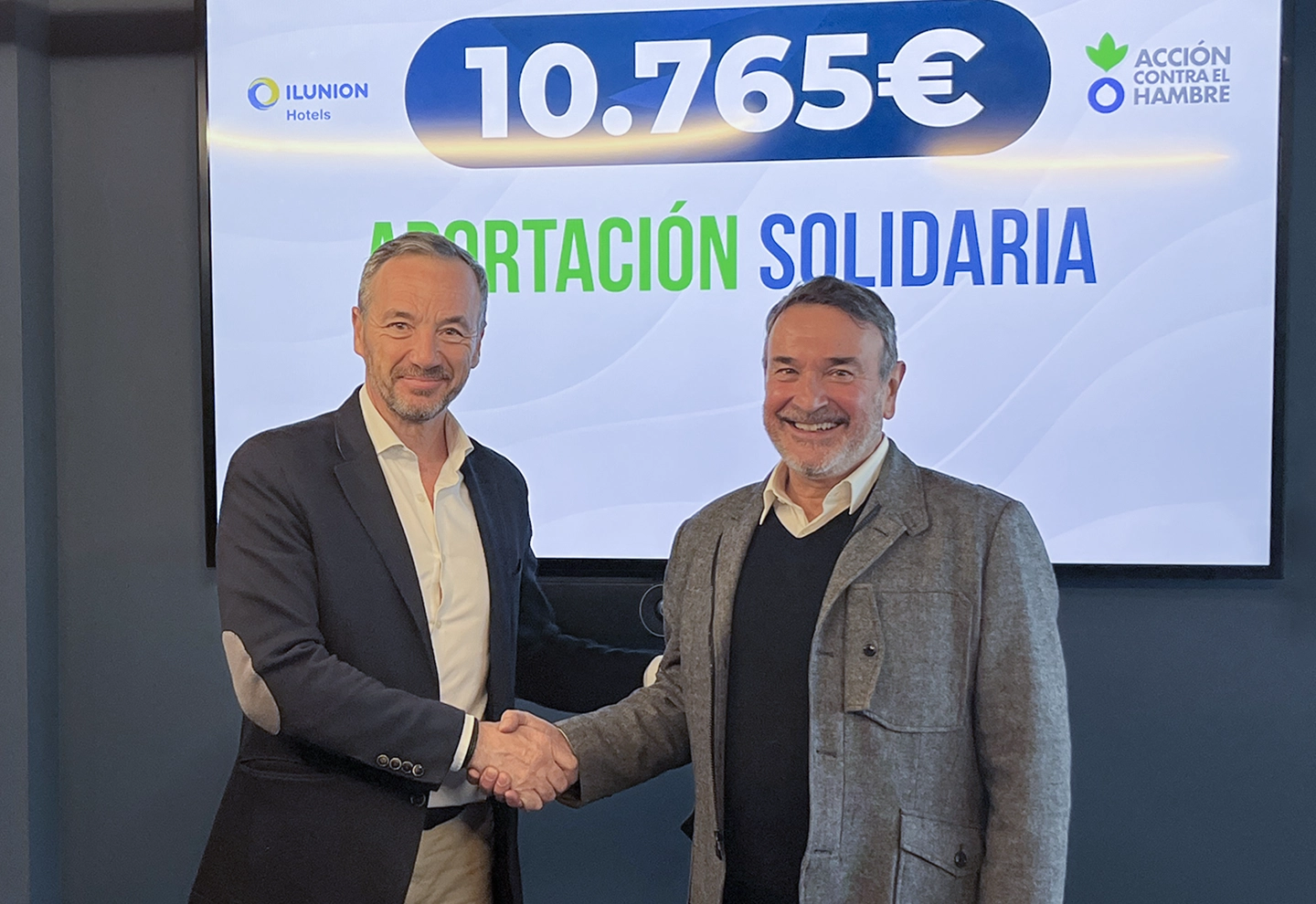 Two people shake hands in front of a screen displaying the text “ILUNION Hotels”, “€10,765” as a charitable contribution, and “ACTION AGAINST HUNGER”.