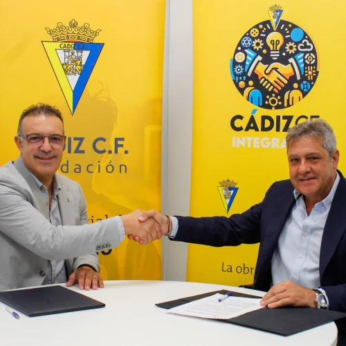 Two men shake hands in a room, seated at a desk, with yellow Fundación Cádiz CF banners behind them.