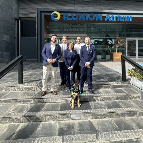 The ILUNION Atrium team poses with Lía, the first guide dog to be raised in a hotel. They stand smiling in front of the hotel entrance on a sunny day.
