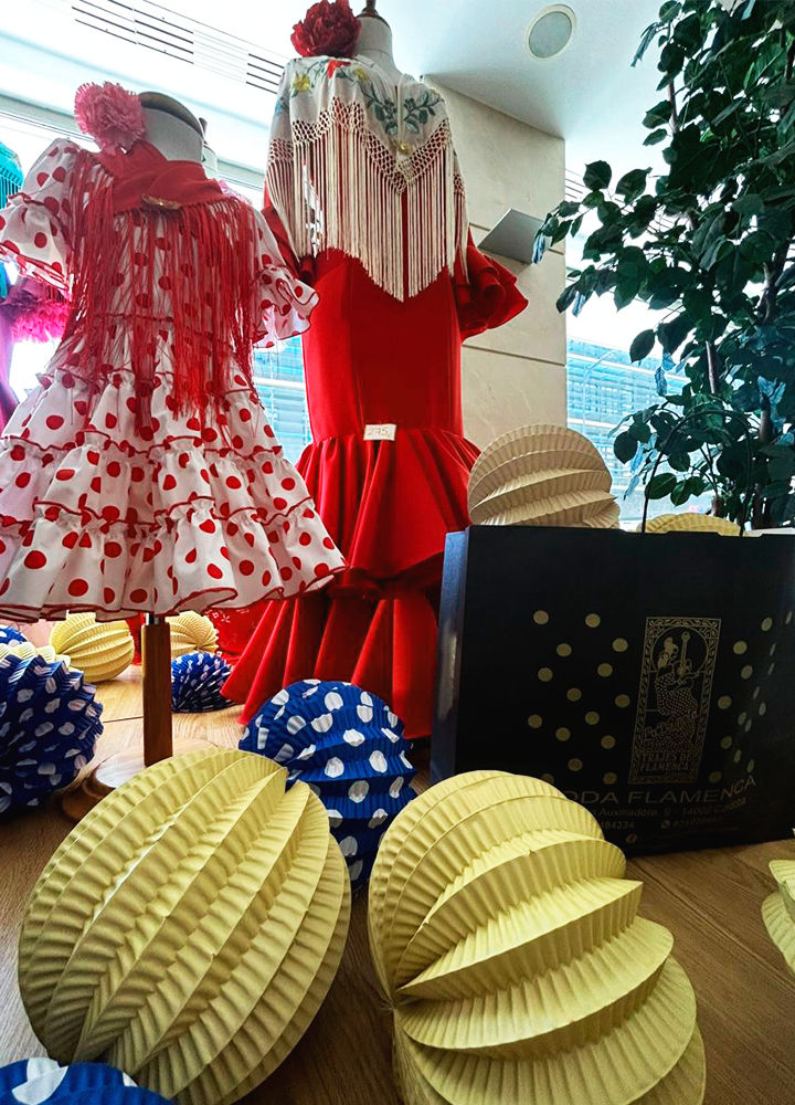 Dos maniquíes visten trajes flamencos con volantes y mantones, rodeados de adornos festivos de papel; uno lleva vestido blanco con lunares rojos y el otro, vestido rojo con mantón bordado, junto a una bolsa negra con texto dorado que dice 