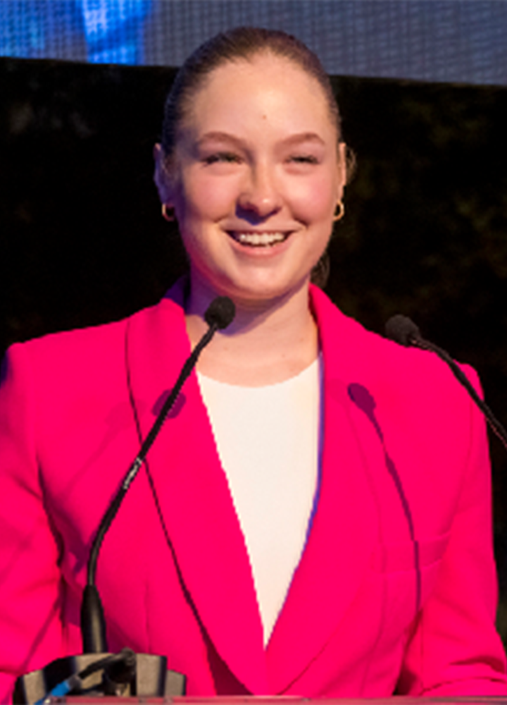 Smiling person wearing a pink blazer and white t-shirt speaks at a public event in front of two microphones.