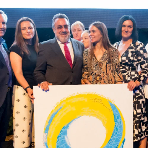 Six elegantly dressed people pose together holding a large circular painting in blue and yellow representing the ILUNION Hotels logo.