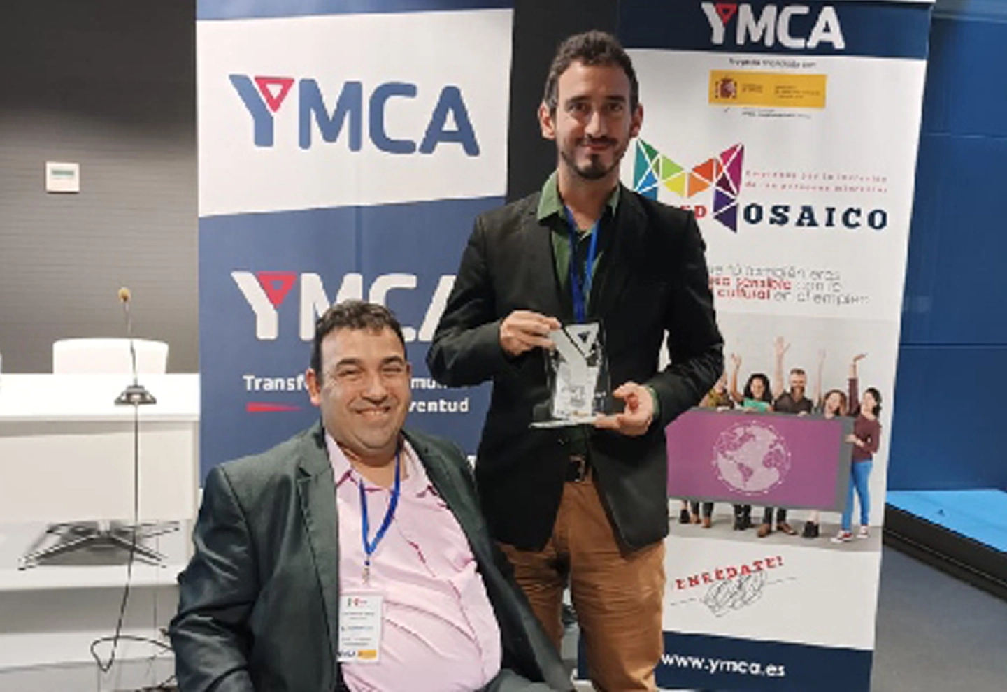 One person stands holding a trophy next to another who is seated, both at an event with banners in the background reading “YMCA” and “MOSAICO.”