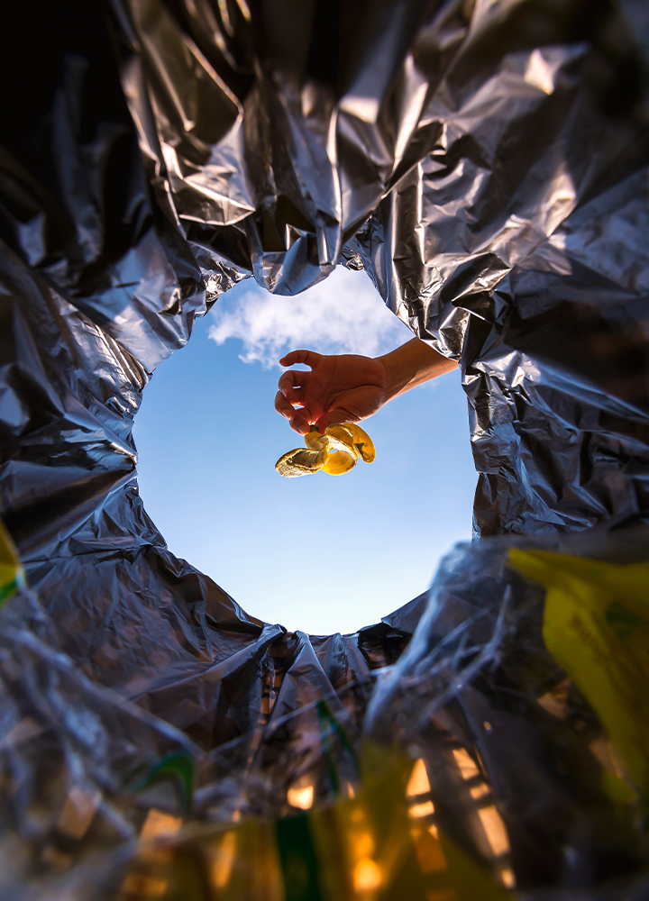 Mano está arrojando una cáscara de plátano a una bolsa de basura negra, con el cielo azul y nubes de fondo.