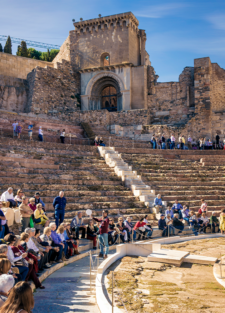 Antiguo anfiteatro romano al aire libre, lleno de turistas sentados en gradas de piedra semicirculares, mientras un guía turístico les explica la historia del lugar bajo un cielo despejado y frente a muros con arcos de piedra desgastados por el tiempo.