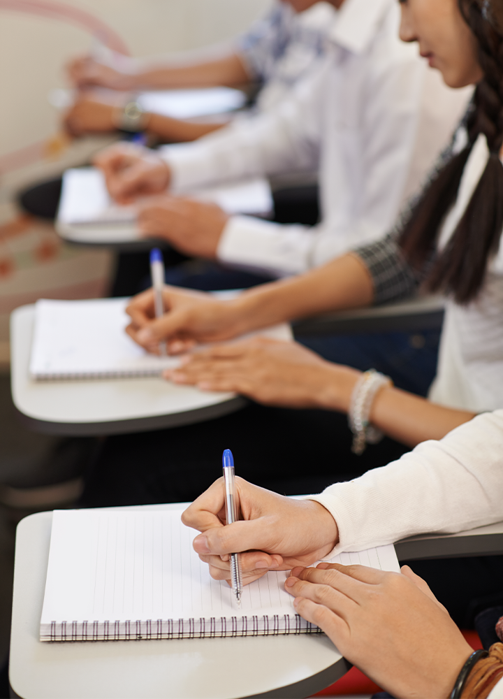 Grupo de personas sentadas en pupitres, escribiendo en cuadernos con bolígrafos.