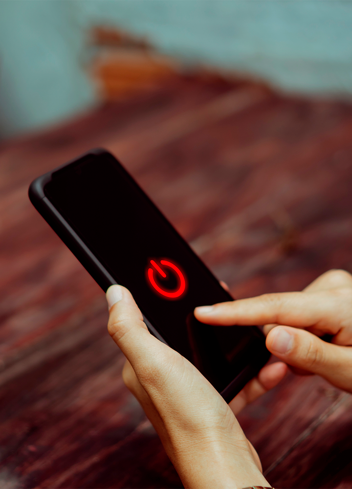 Una mano sostiene un móvil con la pantalla encendida que muestra un gran botón rojo de encendido, mientras otra mano se acerca a tocarlo.