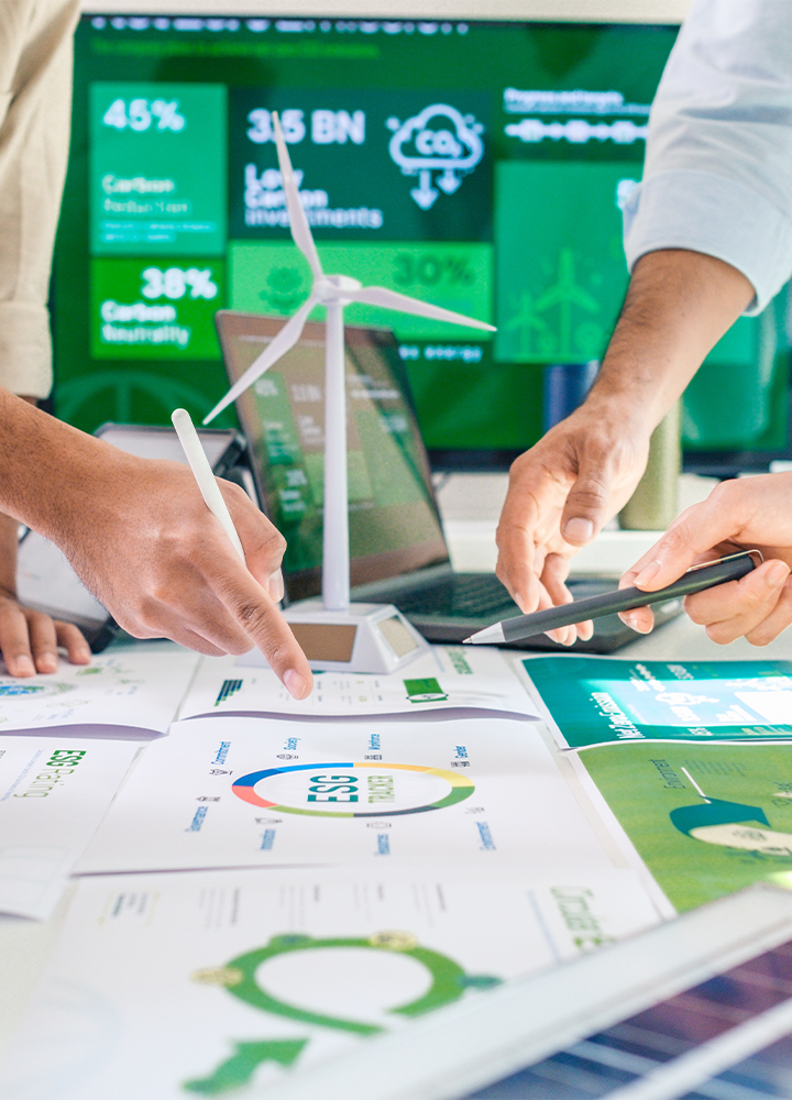 Varias manos manipulando documentos con gráficos ecológicos, una maqueta de turbina eólica y una pantalla que indica datos.