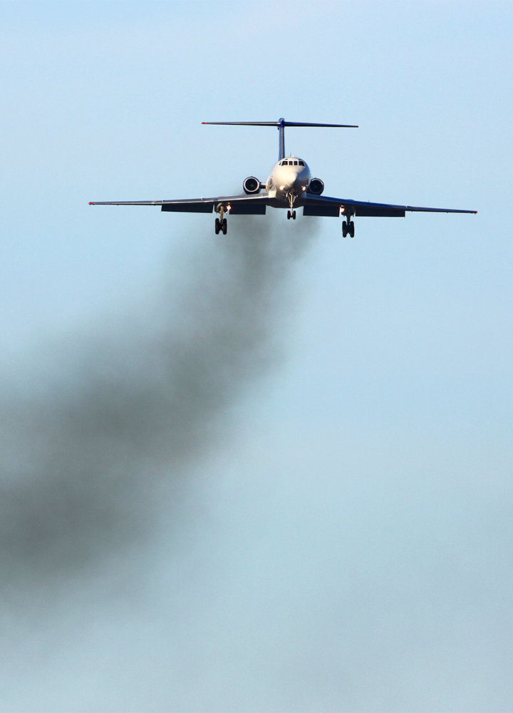 Avión visto de frente vuela dejando una estela de humo negro que contrasta con el cielo azul.