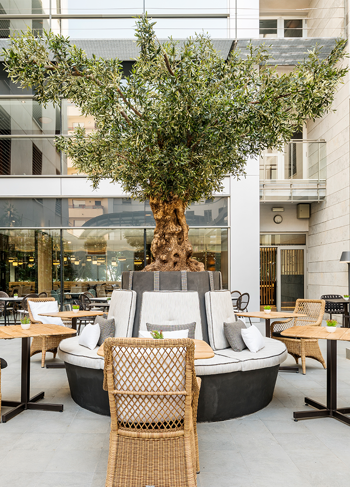 Outdoor terrace with a large tree at the centre, surrounded by circular sofas and tables with chairs.