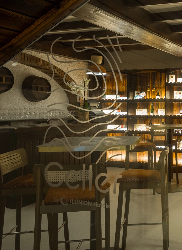 A wine cellar room with high tables and shelves elegantly filled with wine bottles under soft lighting. On the glass, the words “Celler by ILUNION Hotels” can be read.