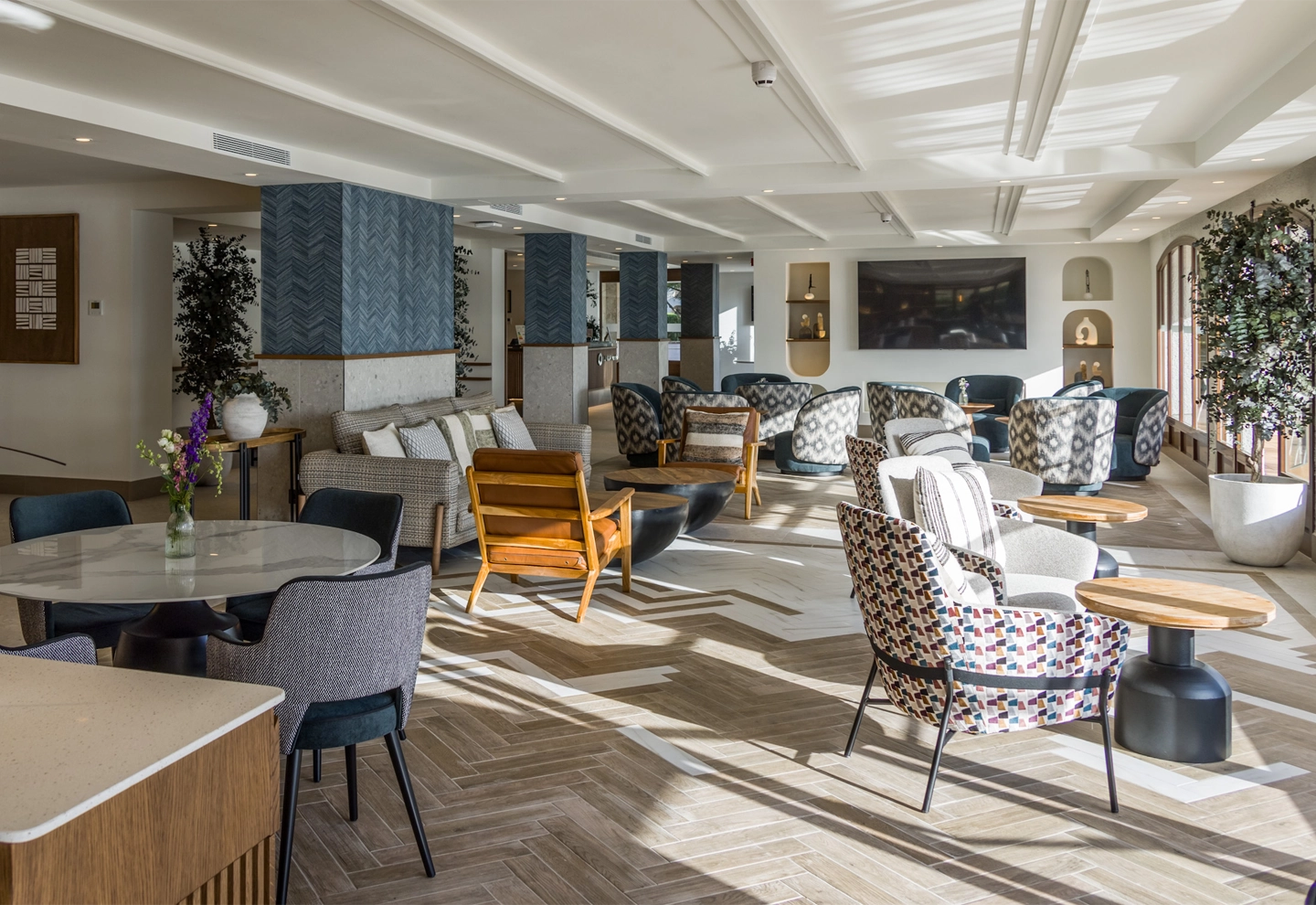 The image shows a modern, spacious, and elegant living room with a variety of seating options. The room features a blend of contemporary and mid-century modern design elements, characterized by clean lines, geometric patterns, and a mix of textures.