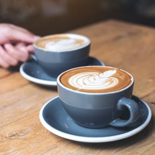 Close-up view of two cups of coffee with milk, both featuring intricate latte art, resting on a rustic wooden table.
