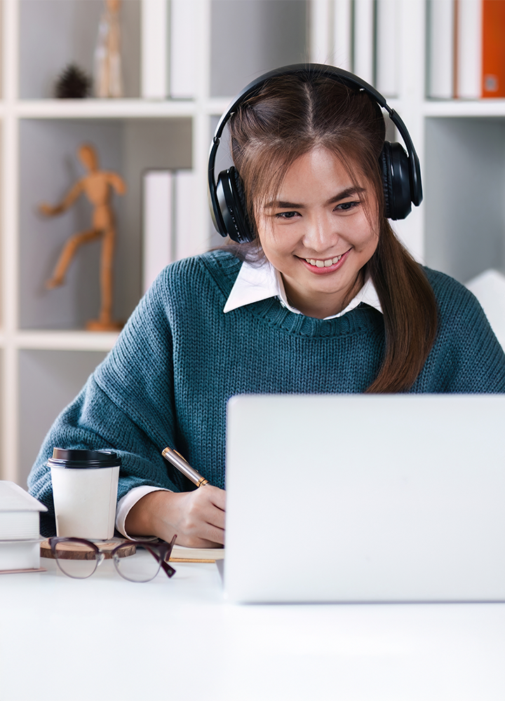 Persona con auriculares trabaja concentrada frente a un ordenador portátil, rodeada de libros abiertos, una taza de café y unas gafas.
