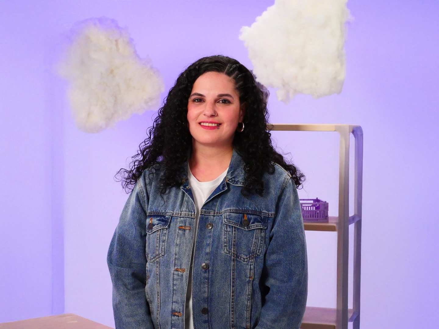 Woman with long, curly hair, wearing a blue denim jacket and a white T-shirt. The background is a soft shade of purple with a few scattered white clouds, evoking a dreamlike or introspective atmosphere.
