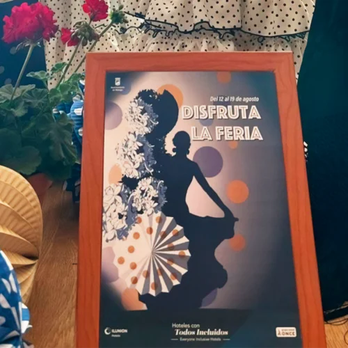 Cartel colorido de feria con una silueta de mujer bailando, flores rojas, abanicos, telas con lunares, y el texto “DISFRUTA LA FERIA” y “Del 12 al 19 de agosto”; aparecen los logos de ILUNION Hoteles y ONCE.