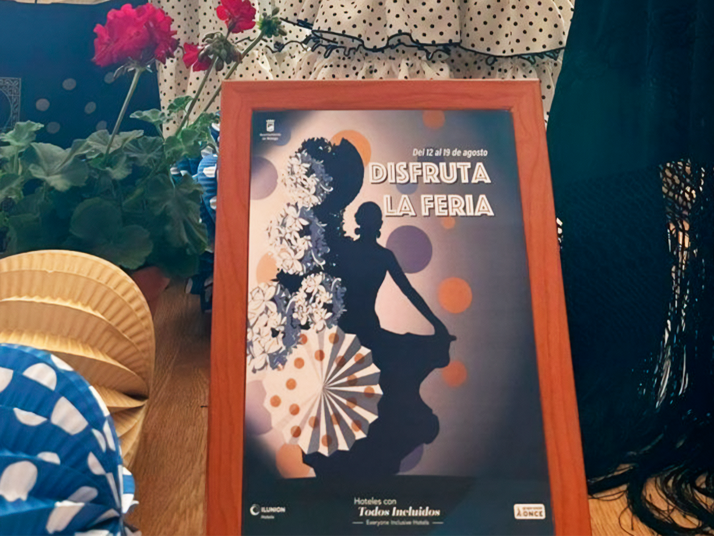 Cartel colorido de feria con una silueta de mujer bailando, flores rojas, abanicos, telas con lunares, y el texto “DISFRUTA LA FERIA” y “Del 12 al 19 de agosto”; aparecen los logos de ILUNION Hoteles y ONCE.