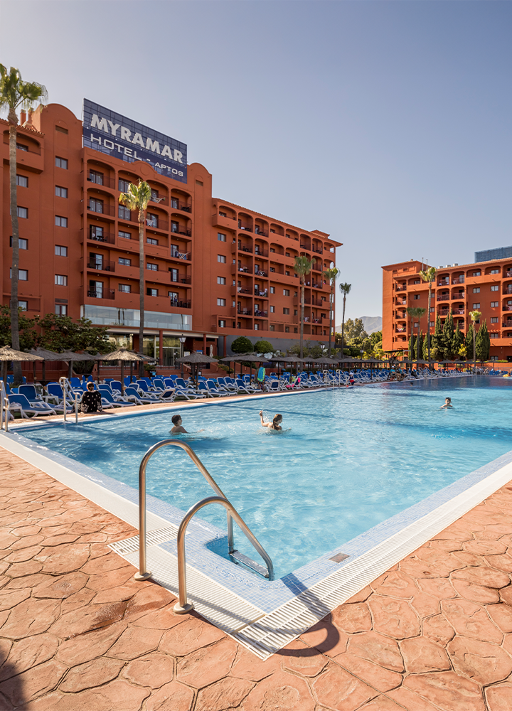 Piscina exterior del Hotel ILUNION Miramar, fachada del edificio cuidada de ladrillo marrón, Hamacas a pie de piscina de color azul. Agua cristalina que refleja un cielo azul despejado.