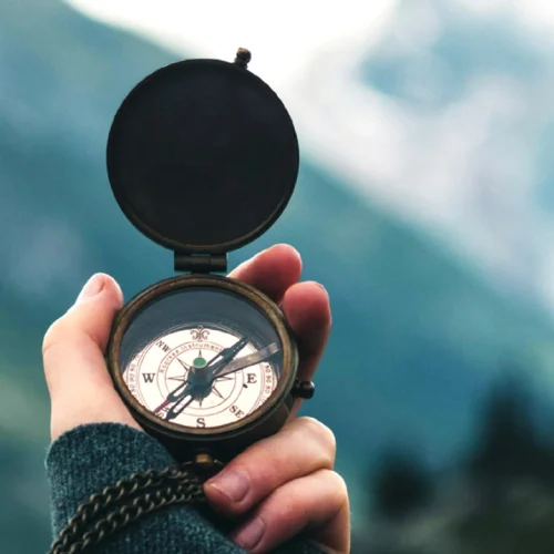 A hand holding a compass pointing north in a mountainous landscape