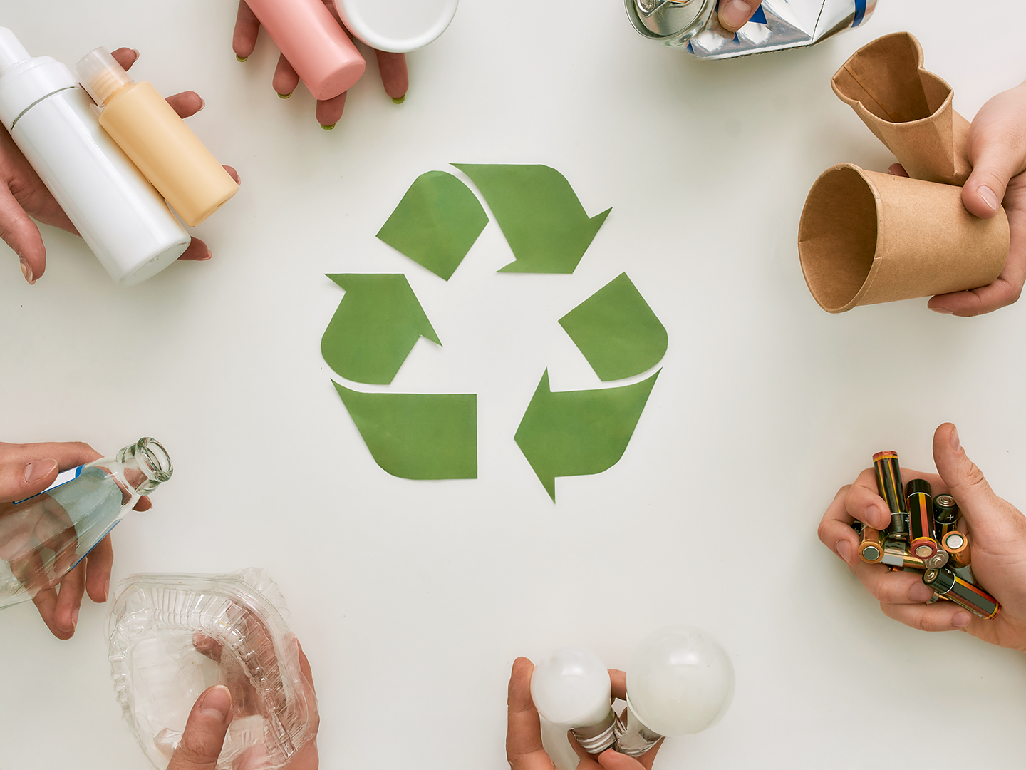 Seis manos sostienen objetos reciclables—botella de vidrio, envase de plástico, lata, batería, bombilla y vaso de papel—alrededor del símbolo verde de reciclaje.