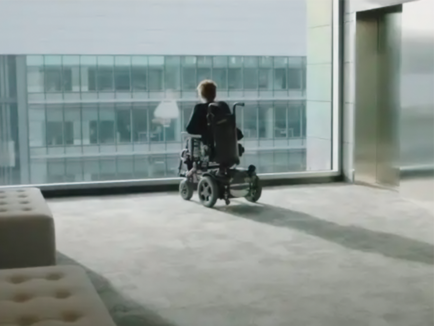 Persona en silla de ruedas viendo el exterior desde una gran ventana acristalada en un edificio moderno.