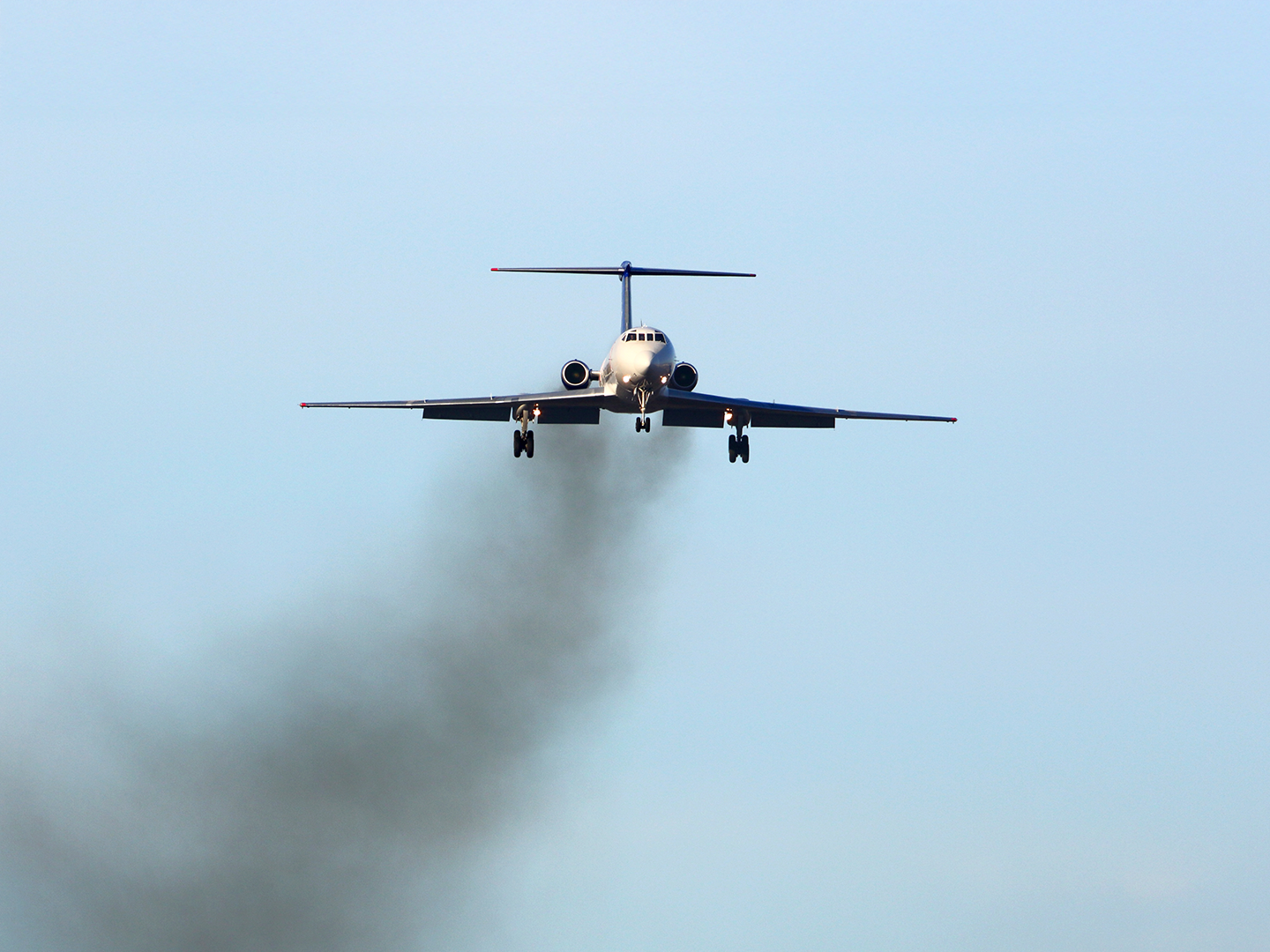 Avión visto de frente vuela dejando una estela de humo negro que contrasta con el cielo azul.