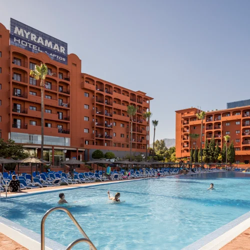 In the photograph, the façade of the ILUNION Miramar hotel can be seen. The façade is brick brown, except for the roofs, which are a darker brown shade. In front of the hotel, there is a large swimming pool with clean, crystal-clear blue water that belongs to the hotel. A cloudless blue sky and sunshine are also visible.