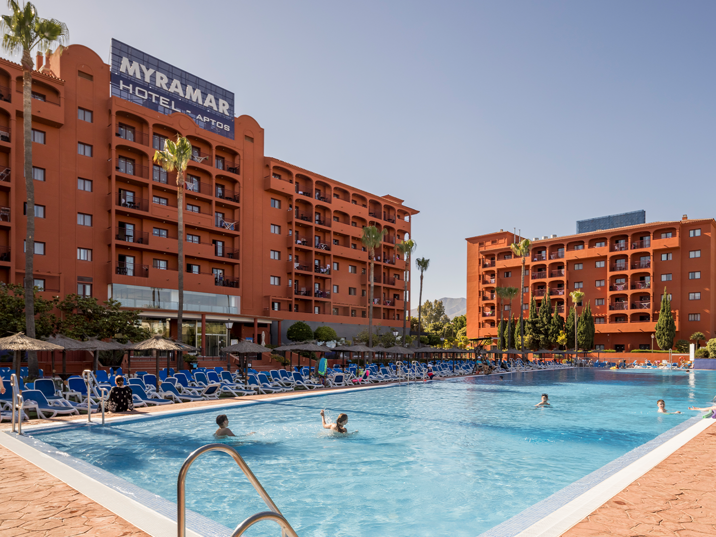 Piscina exterior del Hotel ILUNION Miramar, fachada del edificio cuidada de ladrillo marrón, Hamacas a pie de piscina de color azul. Agua cristalina que refleja un cielo azul despejado.
