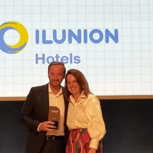 Dos personas están de pie frente a una pantalla; una sostiene un premio, y la pantalla muestra el logotipo circular azul y amarillo junto al texto “ILUNION”