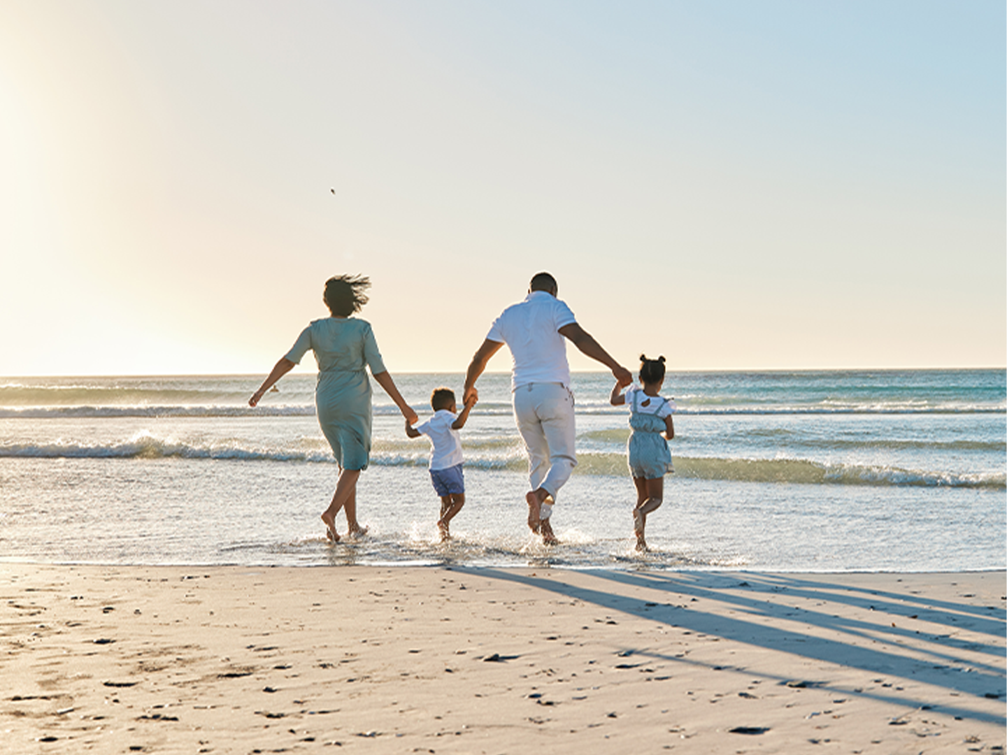 A family of four—two adults and two children—walk hand in hand along the beach toward the sea, illuminated by the soft light of the sunset