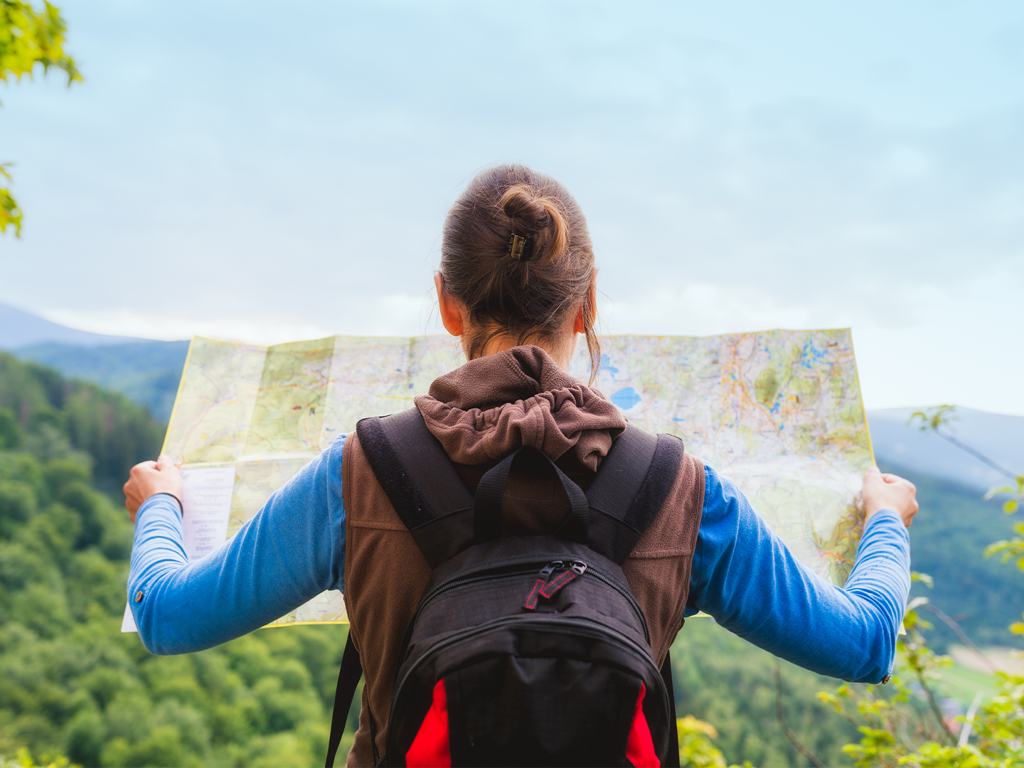 Persona de pie con una mochila en la espalda, en medio de un paisaje verde sostiene un mapa grande con ambas manos,