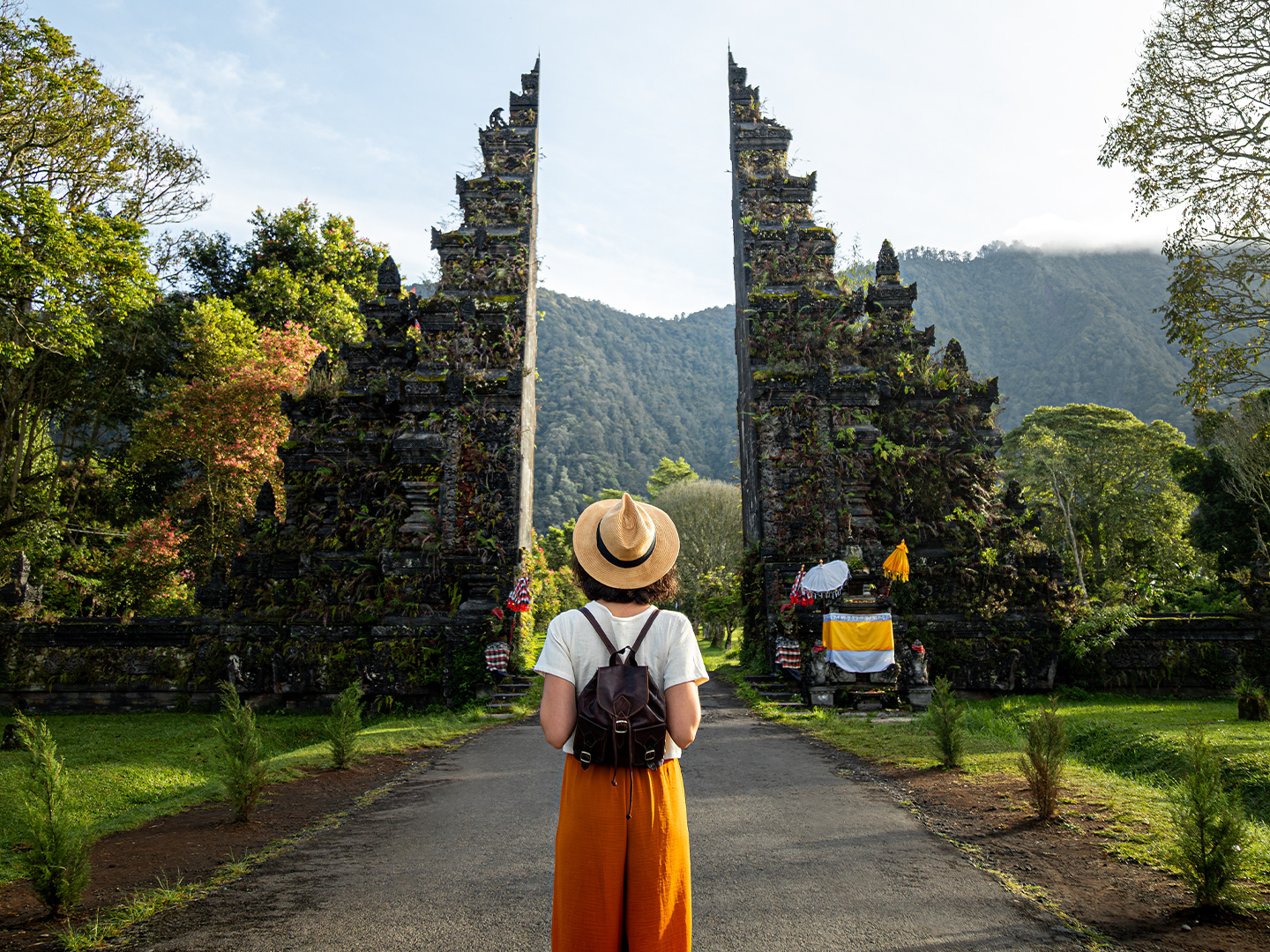 Persona con mochila y sombrero está de espaldas, frente a una monumental puerta balinesa de piedra rodeada de vegetación y montañas.