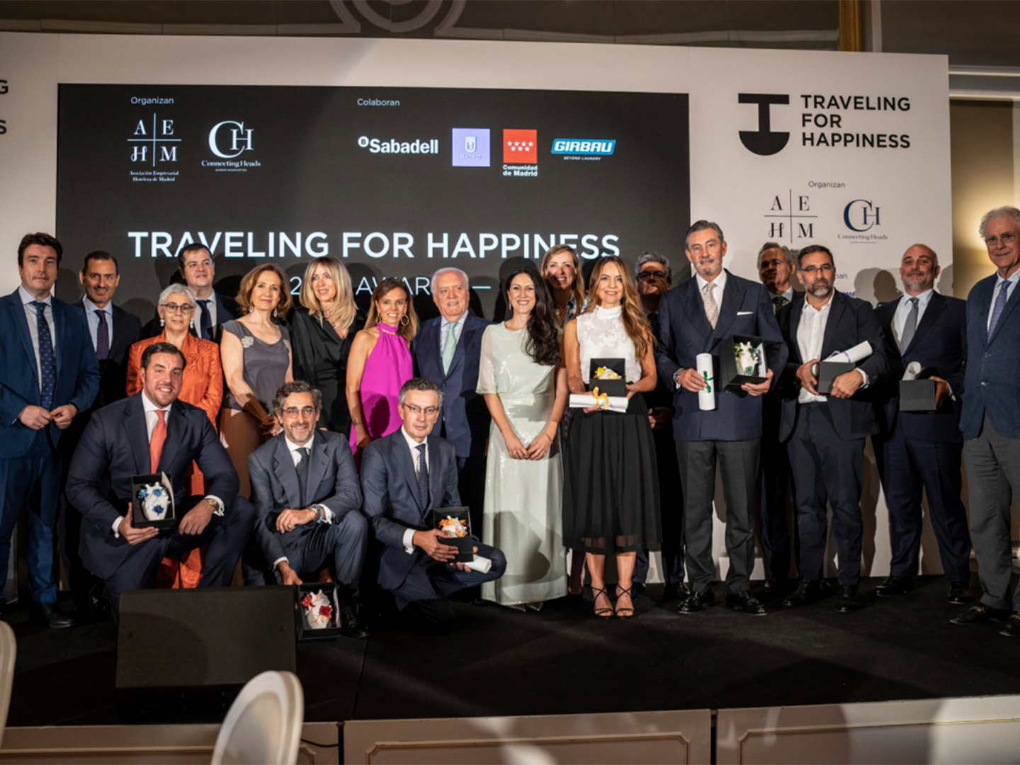 Grupo de personas elegantemente vestidas está de pie en un escenario durante una ceremonia de premios llamada "Traveling for Happiness", con un fondo que muestra logotipos de AEHM, Connecting Heads, Sabadell, Comunidad de Madrid y Girbau.
