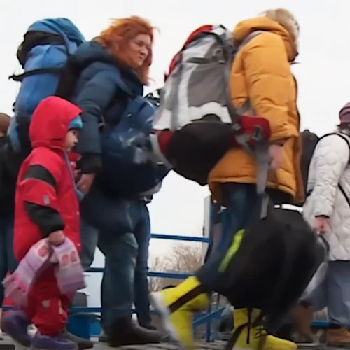 Families and individuals wearing coats and backpacks walk outdoors; a girl dressed in red holds a blanket.