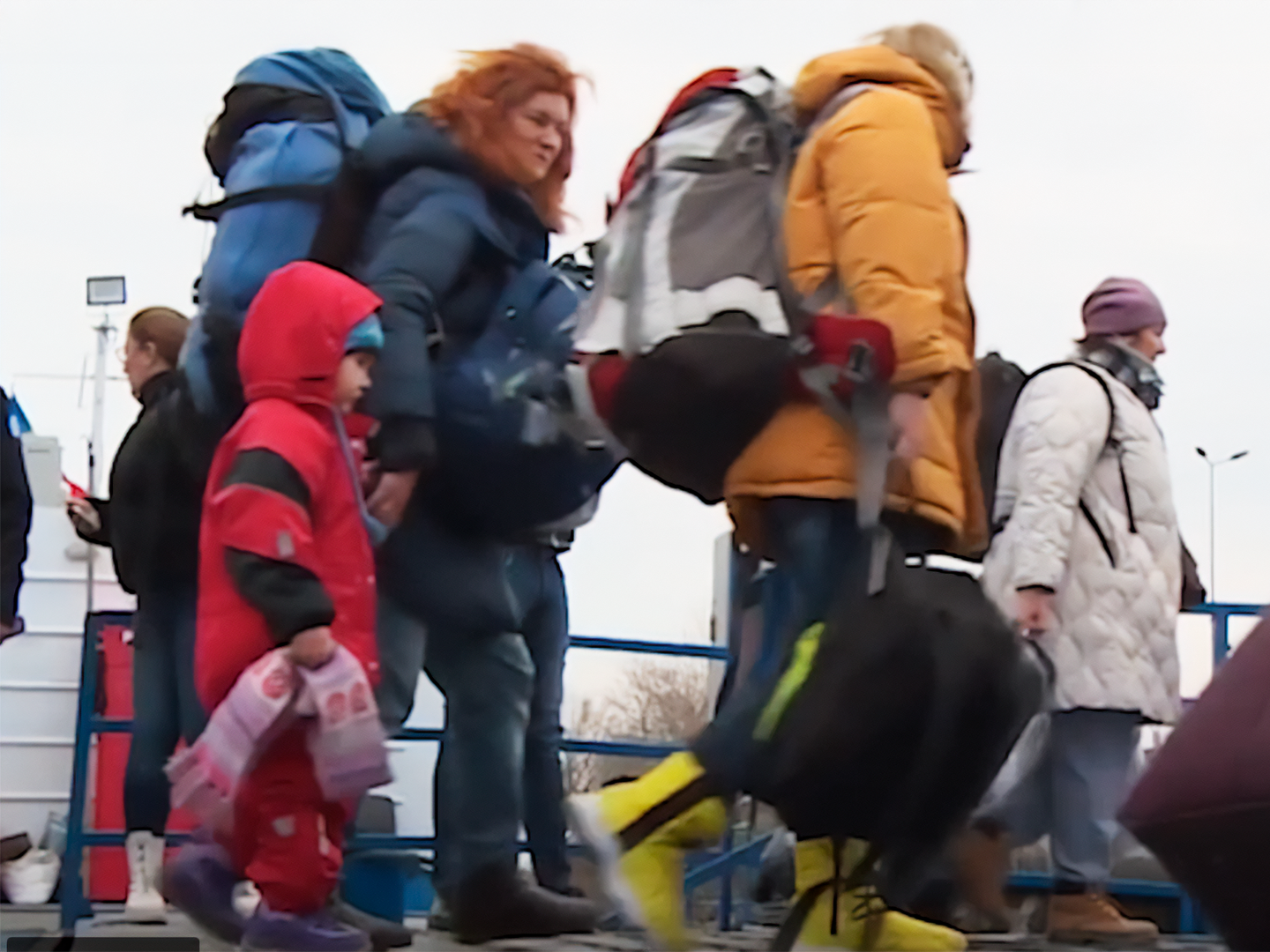 Families and individuals wearing coats and backpacks walk outdoors; a girl dressed in red holds a blanket.