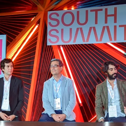 Three people are seated on a stage in front of a colorful backdrop with red, orange, and green lines; above them, a large sign reads “SOUTH SUMMIT.”
