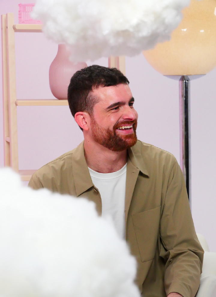 Person wearing a beige jacket over a white shirt in a room with a pastel-colored background. There are various decorative elements, such as a wooden shelf with a pink vase, a large beige-toned lamp, and fluffy white objects hanging from the ceiling, resembling clouds.