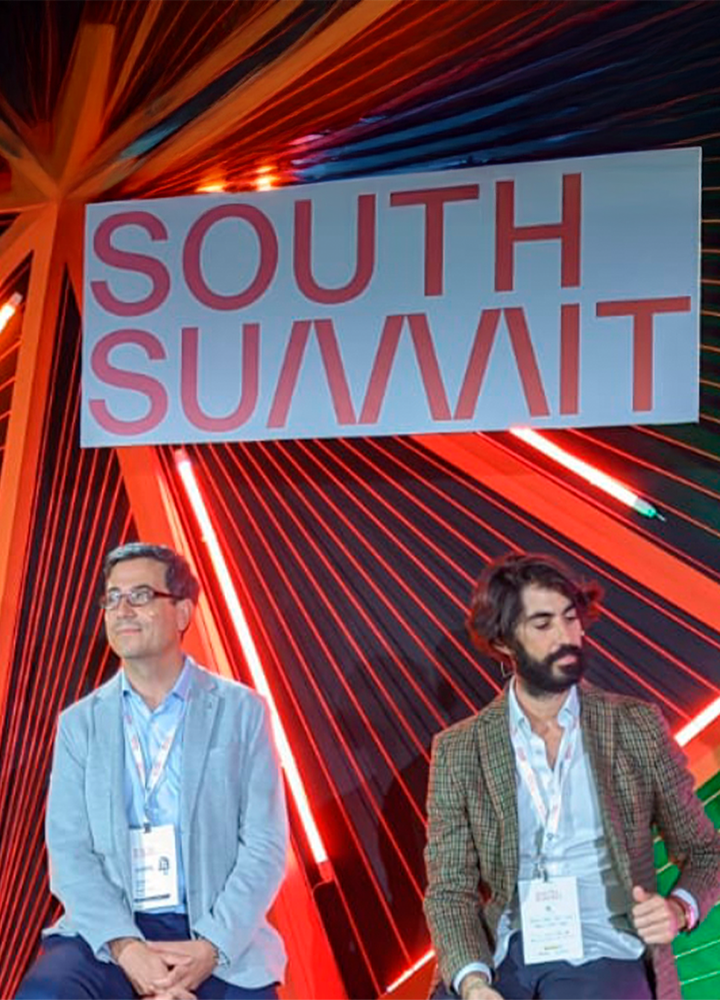 Dos personas están sentadas en un escenario frente a un fondo colorido con líneas rojas, naranjas y verdes; sobre ellos, un gran letrero dice “SOUTH SUMMIT”