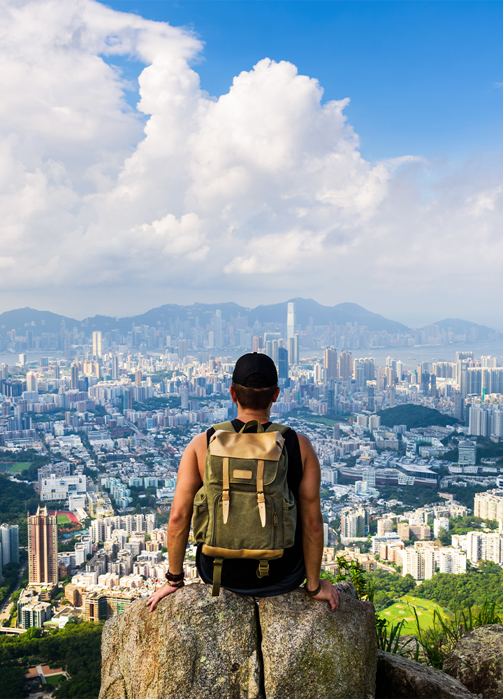 Persona con mochila está sentada en una roca, contemplando una ciudad moderna con rascacielos y montañas en el fondo.