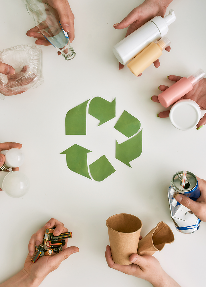 Seis manos sostienen objetos reciclables—botella de vidrio, envase de plástico, lata, batería, bombilla y vaso de papel—alrededor del símbolo verde de reciclaje.