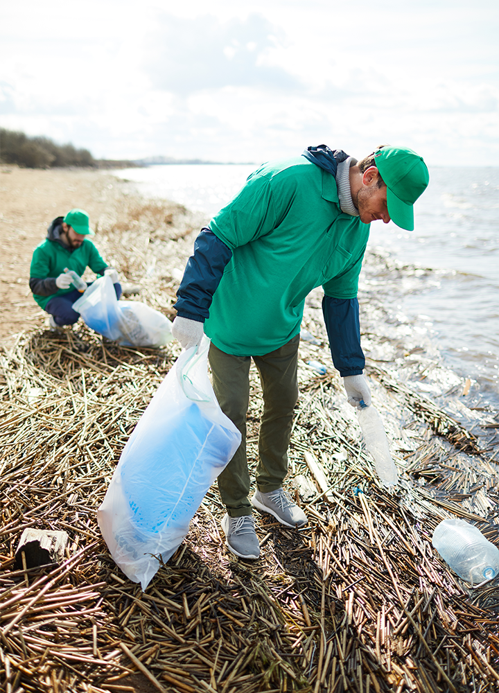 Dos personas con camisetas y gorras verdes recogen basura en una playa.